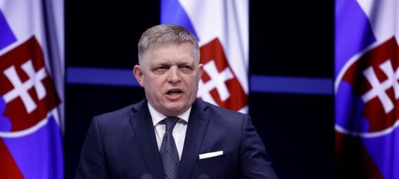 Mężczyzna w garniturze przemawia na tle flag Słowacji