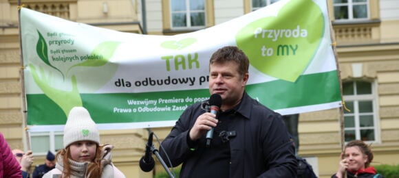 Mężczyzna przemawia na demonstracji, za nim zielone transparenty