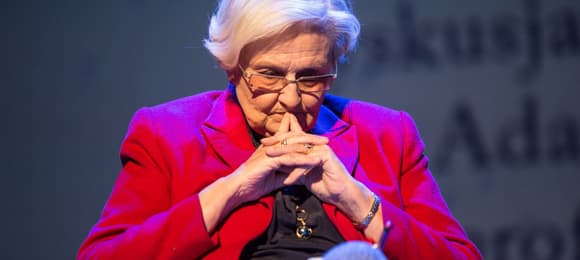 Kobieta w czerwonym żakiecie siedzi w skupieniu trzymająć ręce złożone przy twarzy