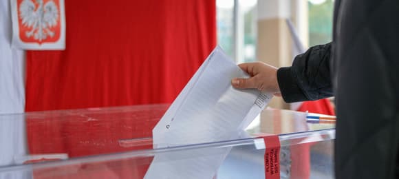 Wyborca wrzuca kartę do głosowania do urny, w tle flaga biało-czerwona i godło państwowe