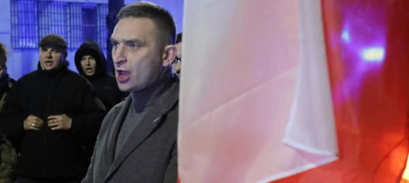 Ubrany w płaszcz mężczyzna krzyczy na ulicy, obok niego polska flaga