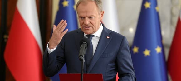 Donald Tusk macha ręką na tle flag Polski i UE stojąc przy mikrofonie