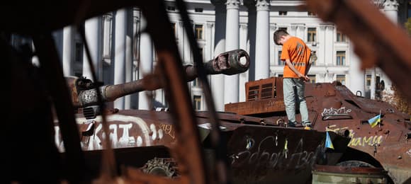 Na zdjeciu widać nastolatka w pomarańczowej koszulce i dżinsach. Stoi tyłem wśród zniszonych czołgów