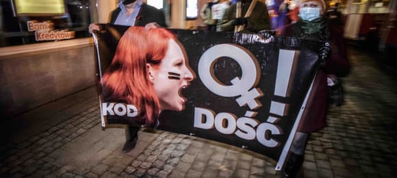 Demonstranci idą ulicą niosąc transparent z rysunkiem krzyczącej kobiety i napisem „Q dość”