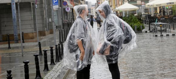 Wrocław, 06.08.2023. Dwóch młodych mężczyzn w pelerynach przeciwdeszczowych stoi w deszczu na wrocławskiej ulicy.