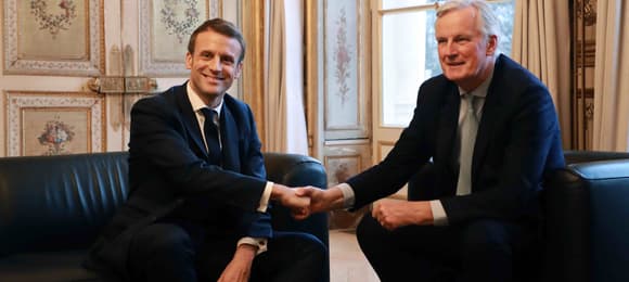 Prezydent Francji Emmanuel Macron ściska rękę z Michelem Barnier , siedząc na kanapie w pałacowej sali