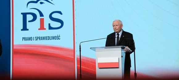 Prezes PiS stoi za mównicą z polską flagą, w tle logo Prawa i Sprawiedliwości