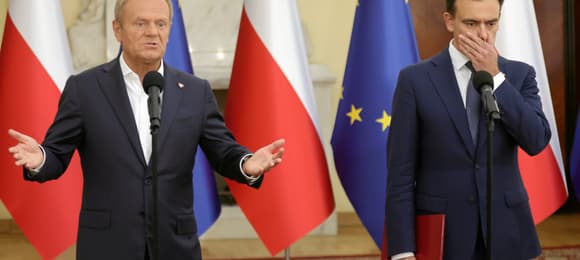 Donald Tusk i Andrzej Domański podczas konferencji prasowej w kancelarii premiera na tle flag Polski i UE, Domański zakrywa część twarzy dłonią