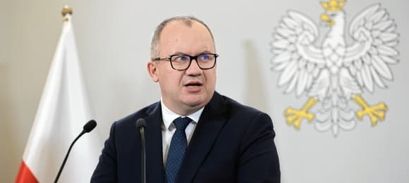 Adam Bodnar na tle biało-czerwonej flagi i polskiego godła. Przemawia do mikrofonów.