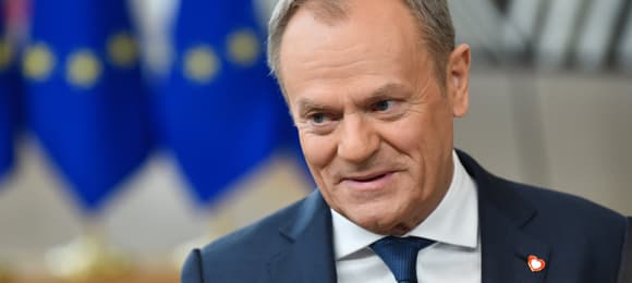 Premier Polski Donald Tusk w niebieskim garniturze i niebieskim krawacie uśmiecha się rozmawiając z dziennikarzami tuż przed rozpoczęciem szczytu Unii Europejskiej w lutym 2024; w tle niebieska flaga UE z żółtymi gwiazdkami