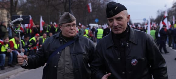 Dwóch mężczyzn w bluzach mundurowych i furażerkach na tle protestujących osób