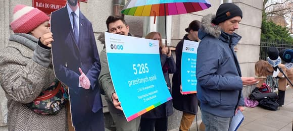 Grupa ludzi przed budynkiem pod tęczową parasolką, z niebieskim plakatem mówiązym, że głosów w konsultacjach ustawy o związkach partnerskich jest 5285 i tekturową figurą wicepremiera Kosiniaka-Kamysza z PSL