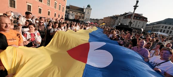 Na Placu Zamkowym w Warszawie tłum rozwija ukraińską flagę. Na środku jest serce w barwach polskiej flagi