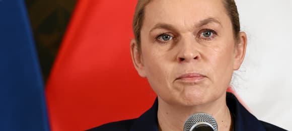 Kobieta (ministra Barbara Nowacka) stoi przed mikrofonem na tle polskiej i europejskiej flagi