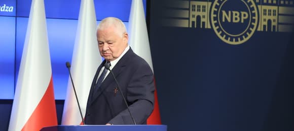 Siwy mężczyzna w garniturze schodzi z mównicy, w tle flagi Polski i logo NBP