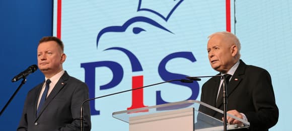 Dwóch mężczyzn, jeden za szklanym pulpitem z biało-czerwoną flagą. Za nimi logo PiS z orłem w koronie. Mariusz Błaszczak i Jarosław Kaczyński