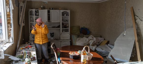 Kobieta w pomarańczowej kurtce stoi w zbombardowanym mieszkaniu.