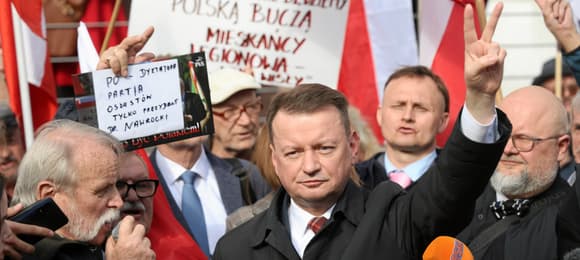 Tłum ludzi z banerami i biało-czerwonymi flagami. Na pierwszym planie Mariusz Błaszczak, mężczyzna z palcami ułożonymi w znak zwycięstwa