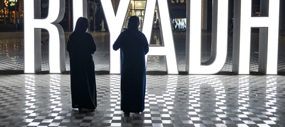 Dwie kobiety w abajach i hidżabach stoją przed wielkim napisem RIYADH.