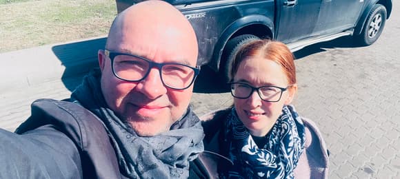 Mężczyzna w okulrach i szczupła kobieta obok, na tle ciemnego samochodu terenowego