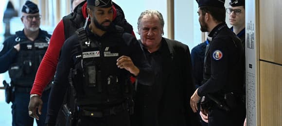 Na zdjęciu widać starszego mężczyznę w ciemnym ubraniu eskortowanego przez kilku francuskich policjantów w mundurach. Znajdują się w jasnym, nowoczesnym korytarzu, prawdopodobnie w budynku sądu. Policjant na przedzie prowadzi grupę, atmosfera jest poważna, sugerująca doprowadzenie na rozprawę sądową.