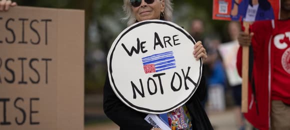 Kobieta trzyma w ręku baner z napisem We are not OK ozdobionym amerykańską flagą