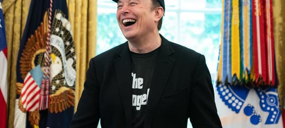 Mężczyzna w czarnym t-shircie, marynarce i czarnej czapce z daszkiem (Elon Musk) donośnie się śmieje. Za nim na ścianie wisi sztandar prezydenta USA (dekoracje Gabinetu Owalnego w Białym Domu)