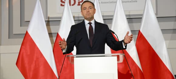 Mężczyzna w ciemnym garniturze (Karol Nawrocki) gestykuluje stojąc przy mównicy na tle biało-czerownych flag