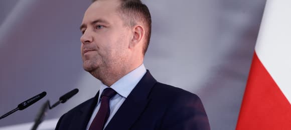 Mężczyzna w średnim wieku, w garniturze, stoi podczas wystąpienia. Z prawej strony fragment biało-czerwonej flagi