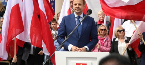 Rafał Trzaskowski za pulpitem z polskim godłem, w tle flagi narodowe