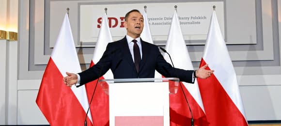 W dużej sali stoi przed mikrofonem mężczyzna i rozkłada szeroko ręce w geście
