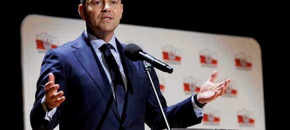 Mężczyzna w średnim wieku w ciemnym garniturze przemawia na mównicy na tle ścianki z biało-czerwonymi symbolami