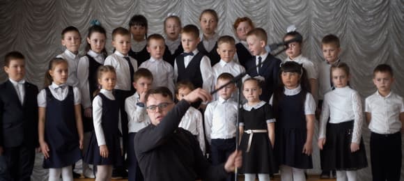 Mężczyzna na pierwszym planie ustawia mikrofon, za nim dziecięcy chór w strojach galowych