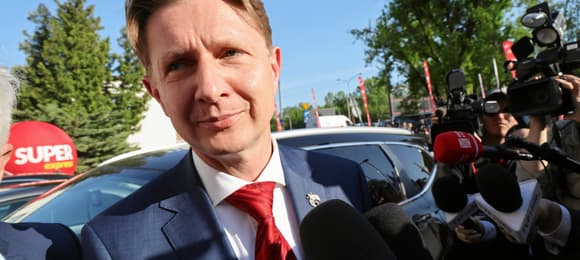Mężczyzna w granatowym garniturze i czerwonym krawacie (Artur Bartoszewicz) uśmiecha się stojąc przed wyciągniętymi ku niemu mikrofonami dziennikarzy