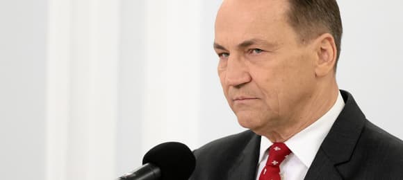 Mężczyzna w ciemnym garniturze w czerwonym krawacie w biały wzór (Radosław Sikorski) stoi przed mikrofonem z poważną miną