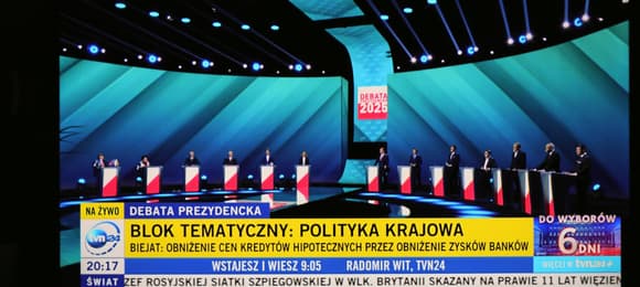 Kadr z debaty prezydenckiej TVP: 13 kandydatów stoi przy pulpitach. Na ekranie napis