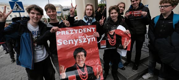 Młodzi ludzie trzymający plakat z Joanną Senyszyn - starszą brunetką w czerwonych koralach.