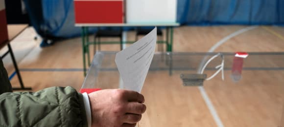 Osoba wrzuca kartę wyborczą do urny, w tle miejsce do głosowania udekorowane białoczerwoną flagą i godłem RP