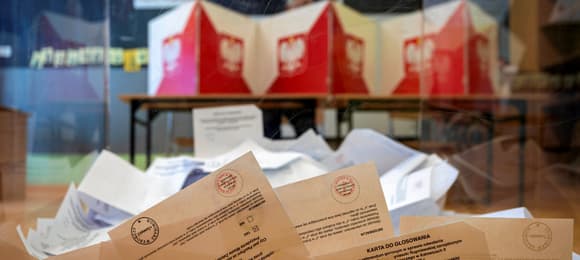 Przezroczysta urna do głosowania, w niej karty do głosowania, w tle kabiny do głosowania z flagą Polski i godłem