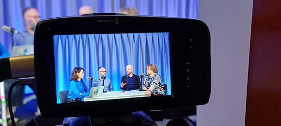 Cztery osoby siedzą przy stole i rozmawiają w studiu nagraniowym. Scena widoczna jest na ekranie kamery Blackmagic Design. W tle niebieska kurtyna, każda osoba ma mikrofon, a jedna z nich ma laptopa z naklejkami.