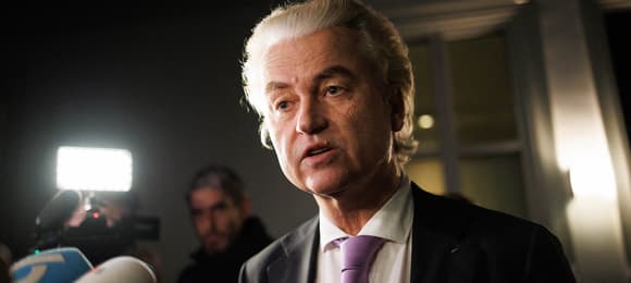 Geert Wilders z Holandii przemawia do mikrofonu. Ubrany w ciemną marynarkę, fioletowy krawat i białą koszulę. Ma zaczesane do tyłu bardzo jasne włosy.