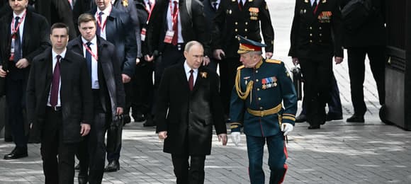 Mężczyzna w czarnym płaszczu (Putin) idzie w towarzystwie bogato udekorowanych orderami wojskowych.