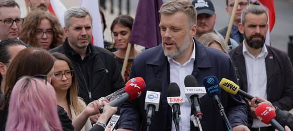 Adrian Zandberg poczas konferencji prasowej w otoczeniu działaczy partii Razem. Przemawia do mikrofonów podstawionych przez media.