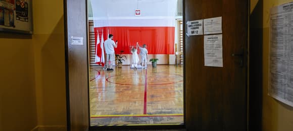 Otwarte drzwi komisji wyborczej, w tle polska flaga i ludzie wrzucający głosy do urny