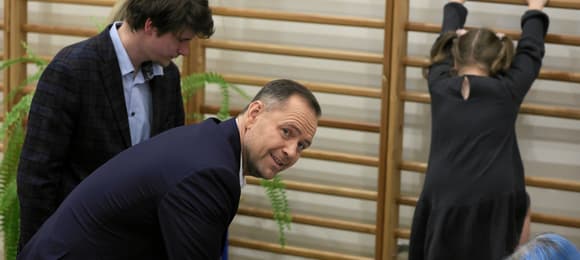 Mężczyzna pochylony nad stołem w lokalu wyborczym, w tle drabinki gimnastyczne, na ktorych bawi się dziewczynka