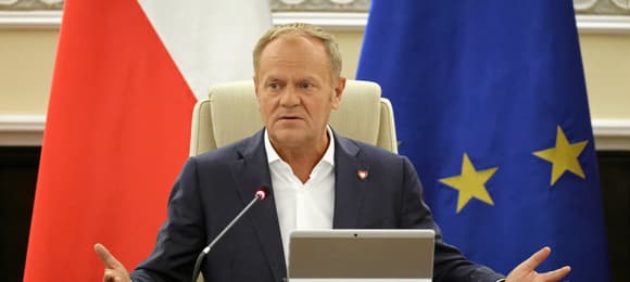 Premier Donald Tusk w sali posiedzeń Rady Ministrów. Siedzi za stołem na tle polskiej i unijnej flagi.