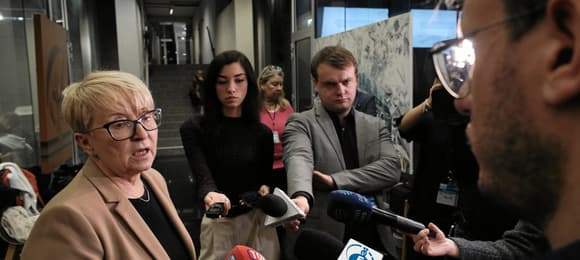 Kobieta, blondynka, krótko ostrzyżona, w okularach odpowiada na pytanie dziennikarzy