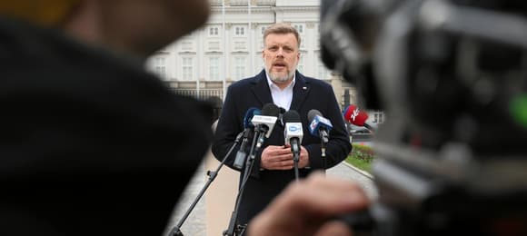 Mężczyzna w płaszczu stoi w tle kadru, zanim Pałac Prezydencki. Na pierwszym planie po obu stronach kamerzyści