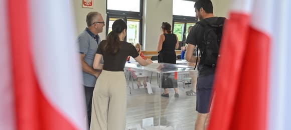 Kobieta w jasnych spodniach wrzuca głos do urny wyborczej, na pierwszym planie po obu stronach kadru polskie flagi