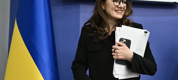 Julia Swyrydenko - kobieta w okularach i garniturze trzymając w ręce plik dokumentów i telefon komórkowy pozuje na tle flagi Ukrainy